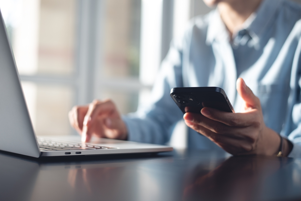 A person in a blue shirt uses a laptop with one hand while holding a smartphone in the other. Indoors, soft daylight filters through windows. The focus is on their hands and devices.