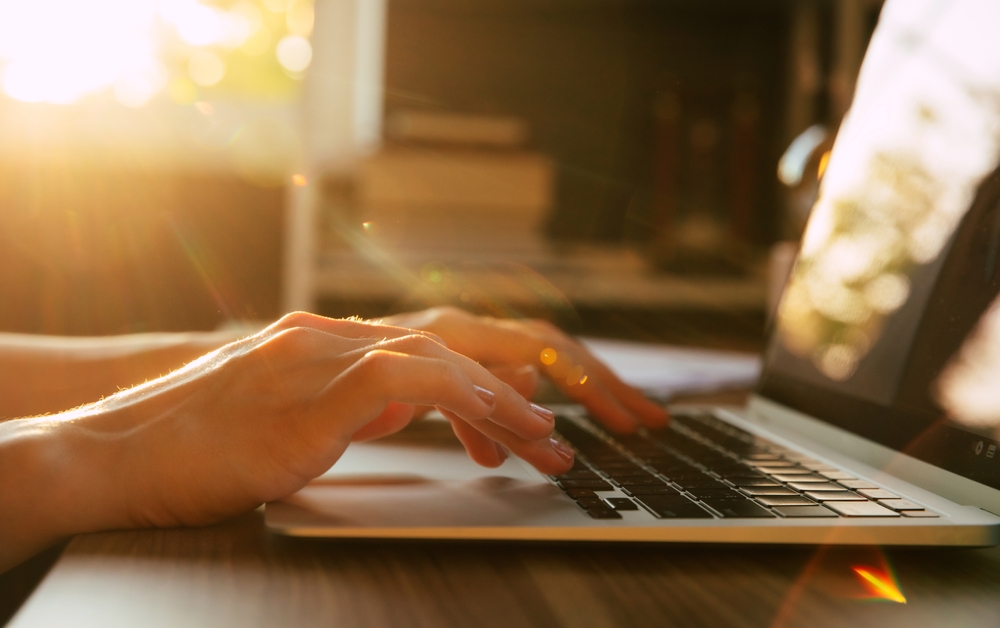 Hands typing on a laptop keyboard on a wooden desk, warm sunlight streaming in from the left. Blurred background with books and shelves creates a cozy, focused workspace atmosphere.