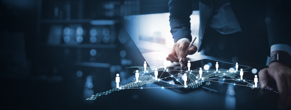 A hand uses a stylus on a tablet displaying digital icons of people connected by lines, symbolizing networking or data connections, with a blurred office background. The image represents technology and business collaboration.