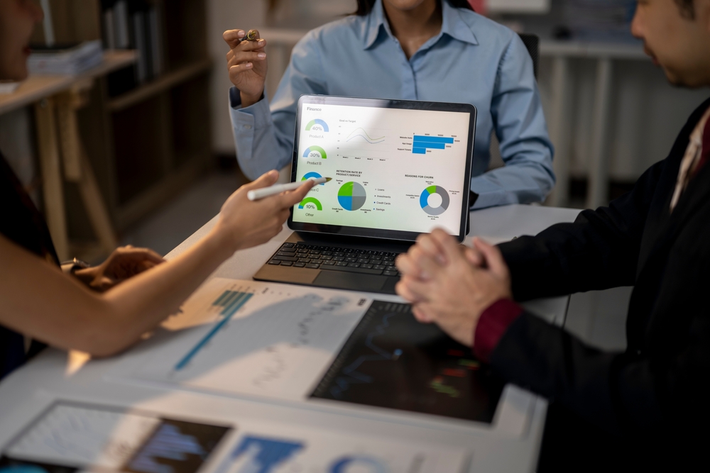 Three people sit around a table with a laptop showing colorful financial graphs and charts. One person points at the screen with a pen. Papers featuring more charts are on the table. Faces are not clearly visible.