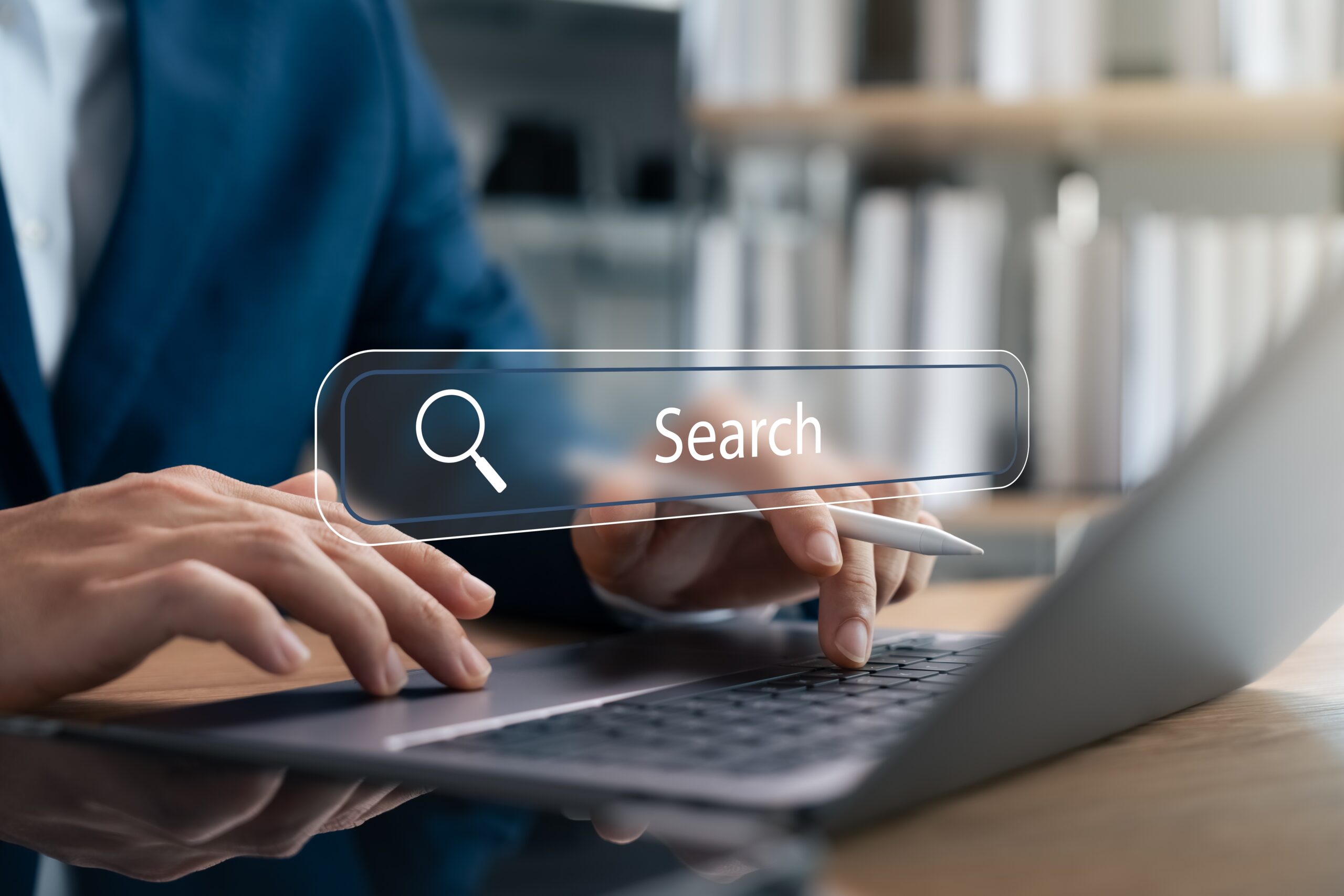A person’s hands typing on a laptop keyboard, one holding a pen. A floating digital search bar with a magnifying glass icon and the word Search appears in front. Blurred bookshelves are visible in the background.