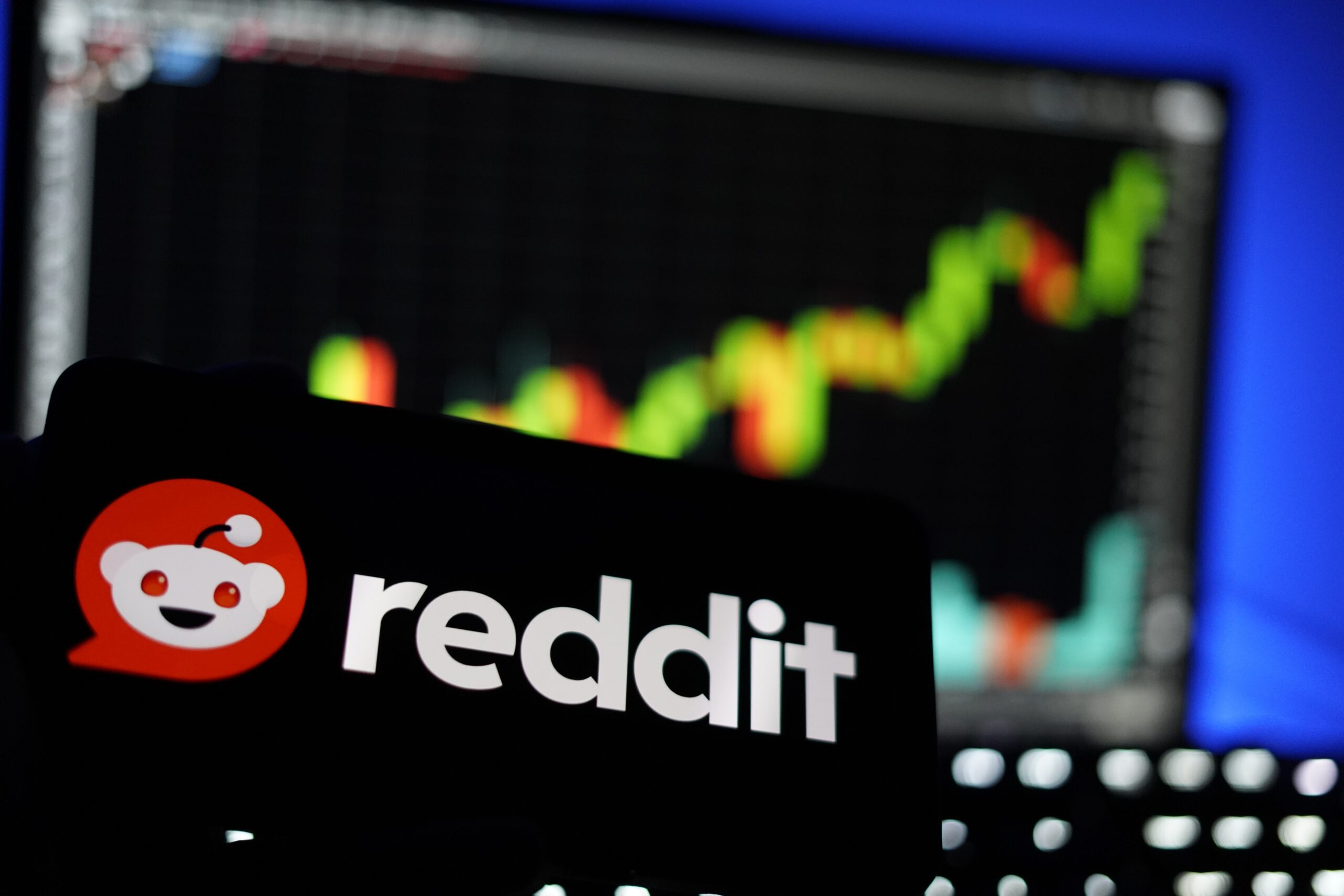 A smartphone shows the Reddit logo in white text on a black background. In the background, a colorful and blurry stock market candlestick chart is visible on a monitor.