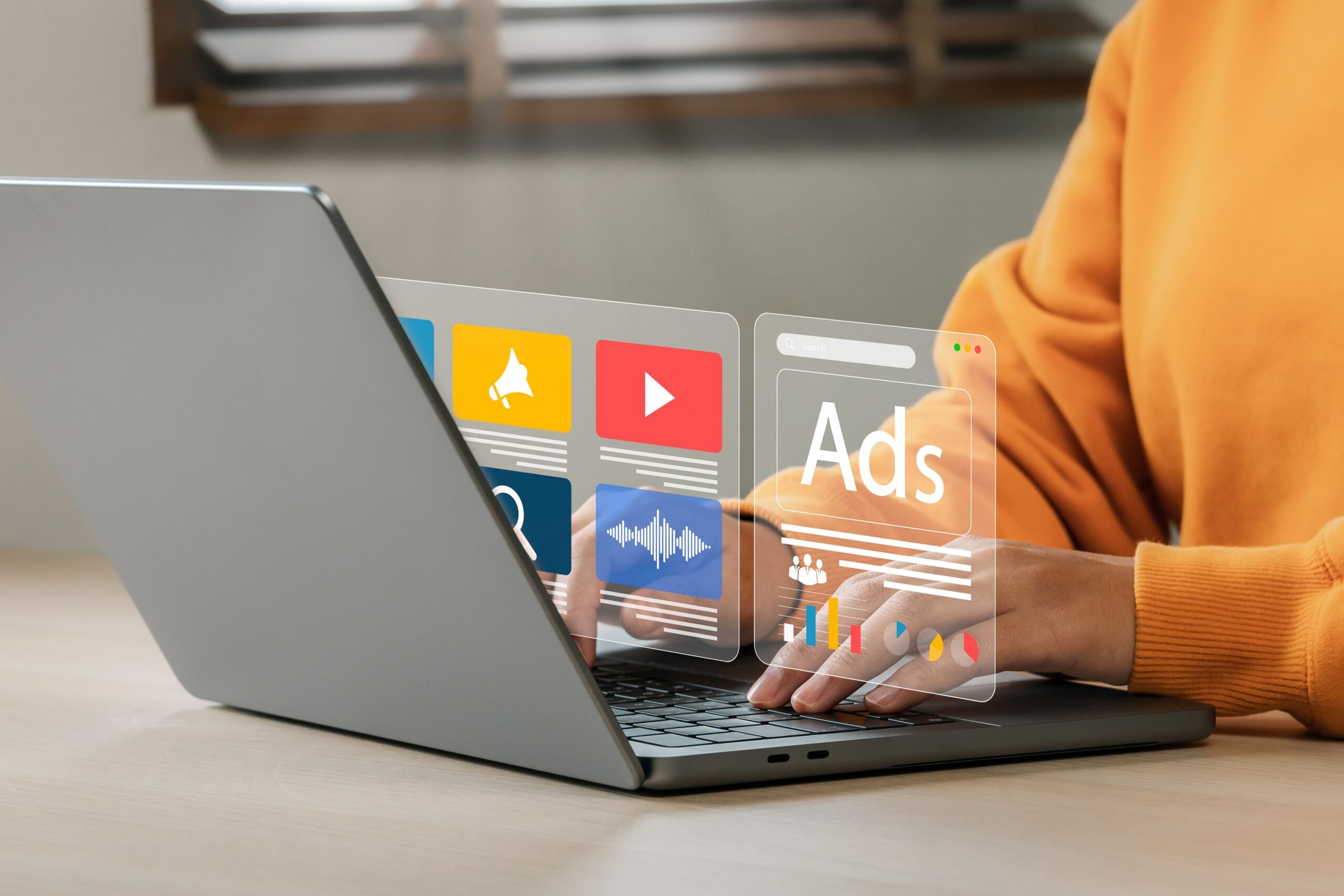 A person types on a laptop while transparent digital icons and an ad management interface float above the keyboard. The interface features various media icons, a bar chart, and the word Ads in large text.